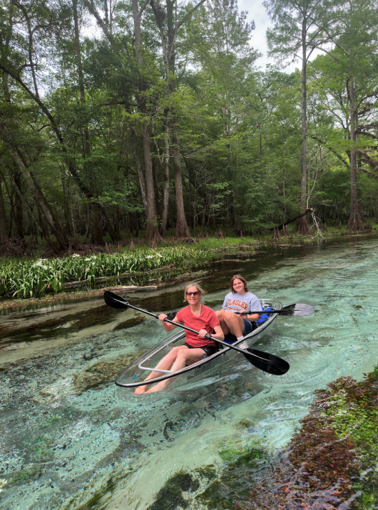 Two people kayaking in a clear boat on a serene, tree-lined river.