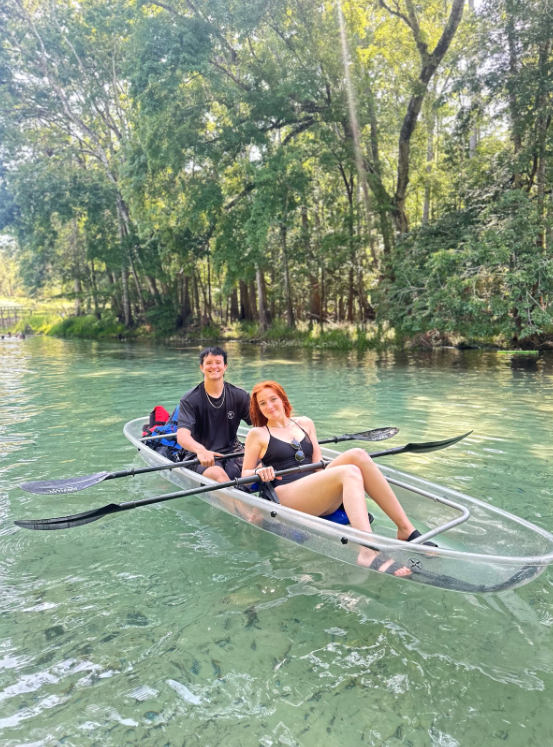 Two people kayaking in a clear kayak on a tranquil river, surrounded by lush green trees.