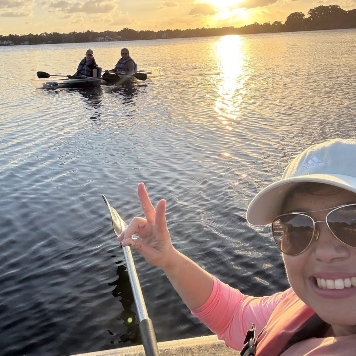 Woman in kayak taking selfie with two people in another kayak, sunset over lake.