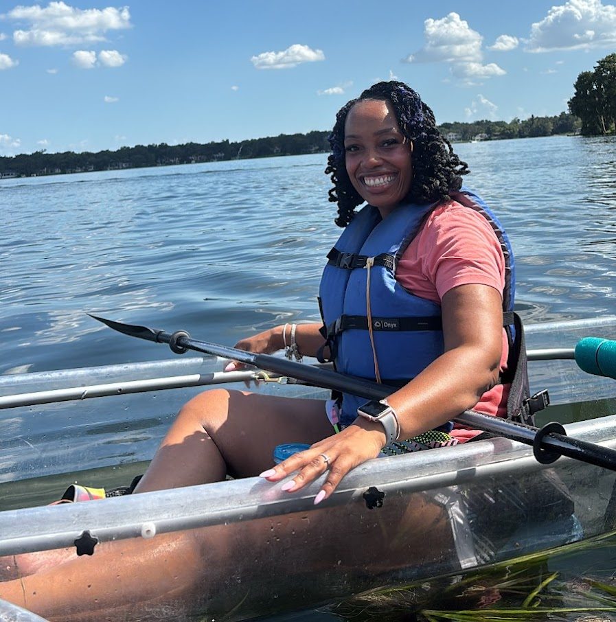 Smiling person in a clear kayak on a lake under a blue sky with fluffy clouds.