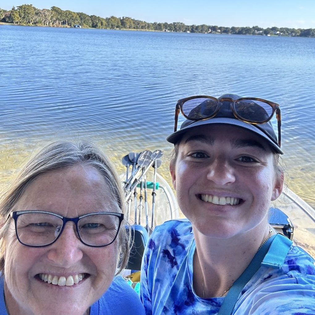 Two people smiling in front of a lake with blue sky and trees.