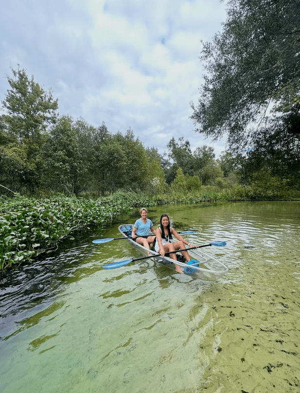 Two people kayaking on a clear river surrounded by trees and greenery under a cloudy sky.