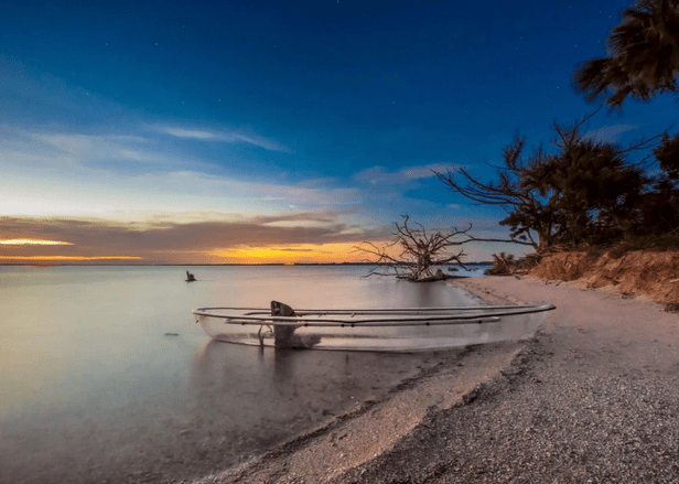 Clear kayak on a sandy beach at sunset with trees and calm water.