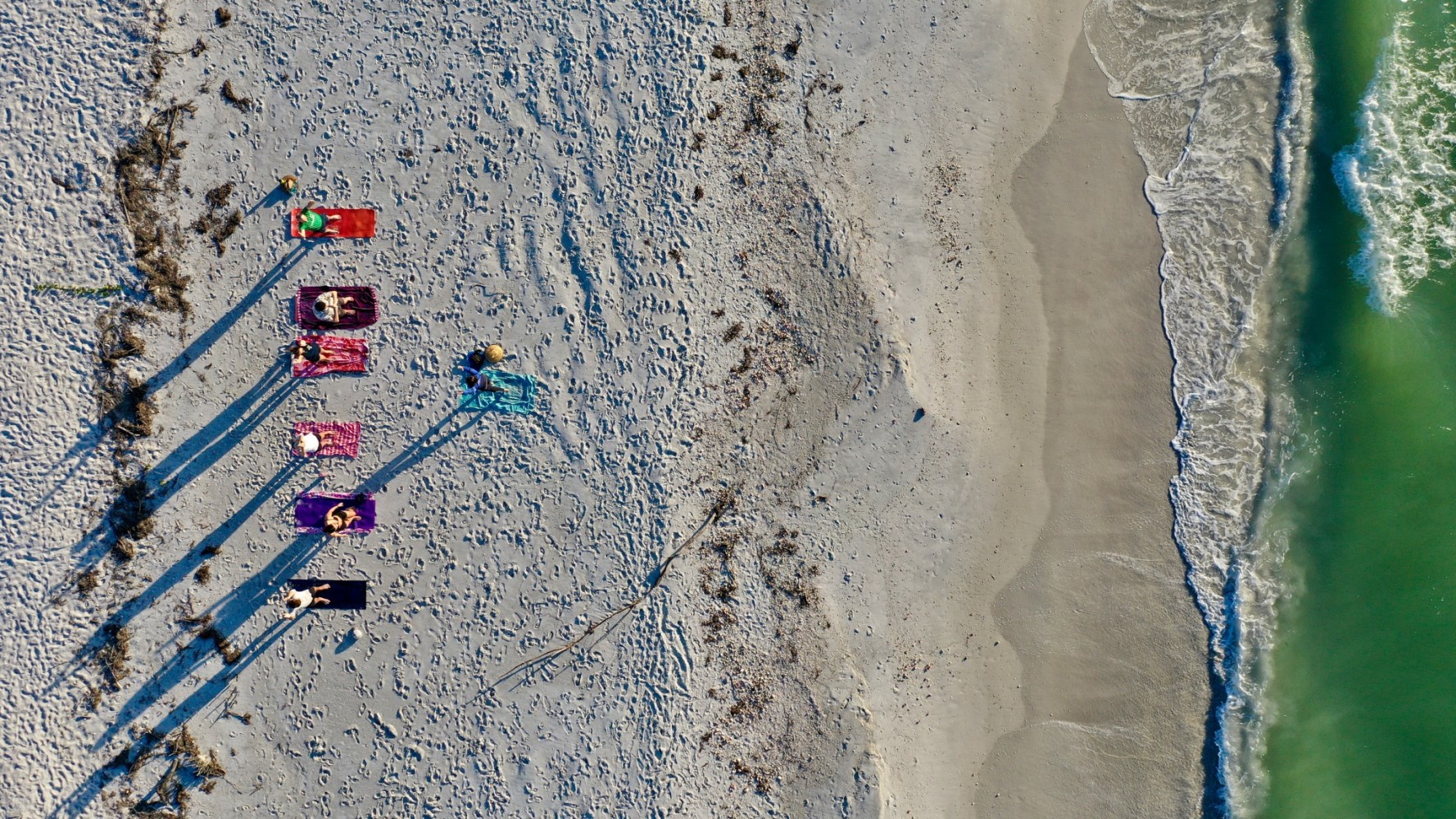 Six people on colorful mats on a sandy beach with ocean waves.