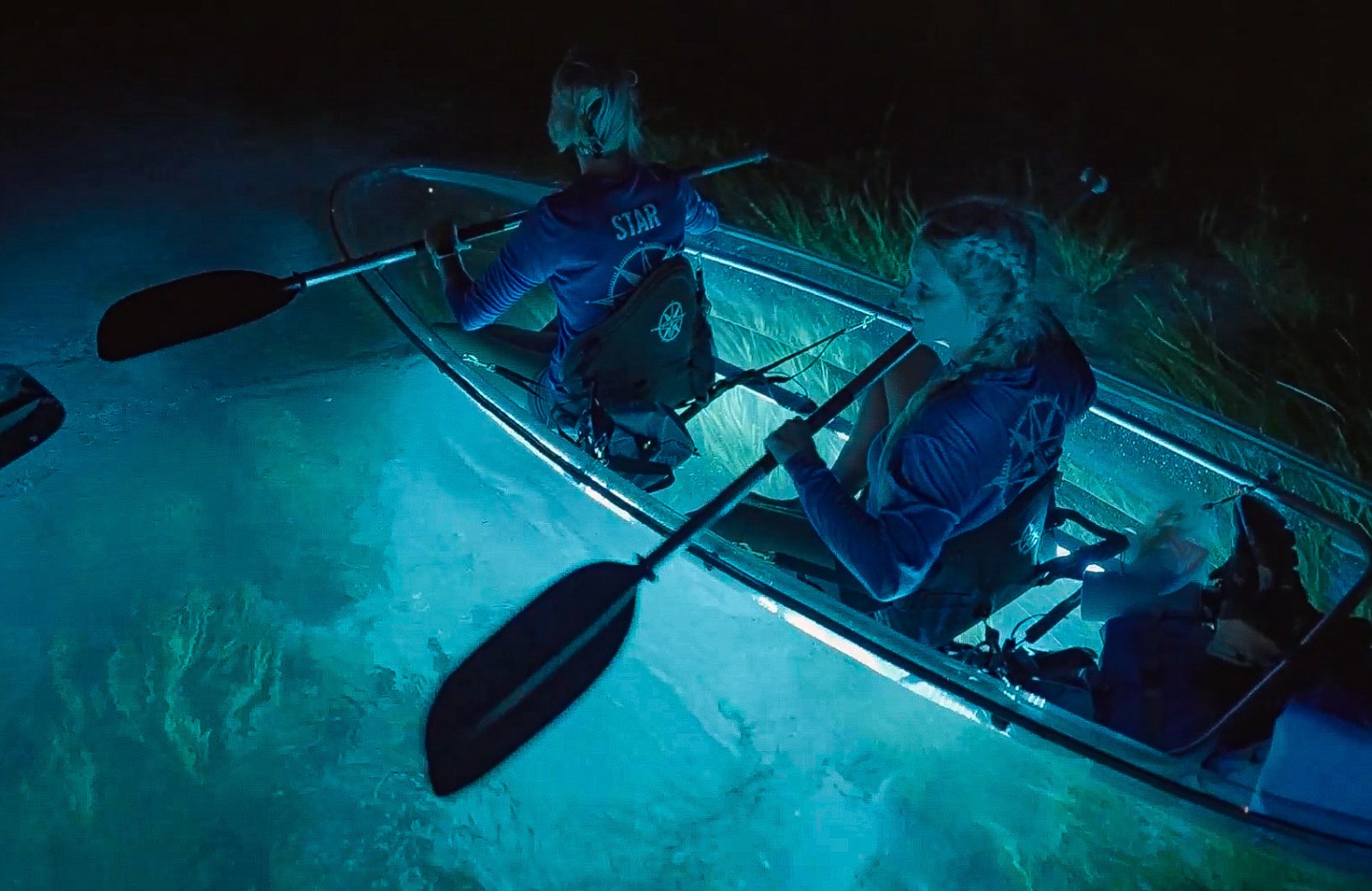 Two people kayaking in a clear-bottom kayak over glowing blue waters at night.
