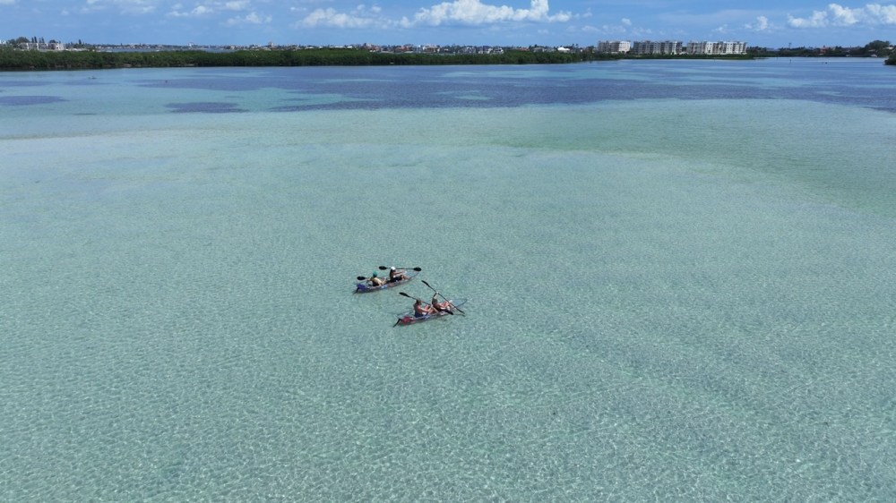Two kayaks with people paddling on clear, shallow turquoise water, with distant shore and buildings.