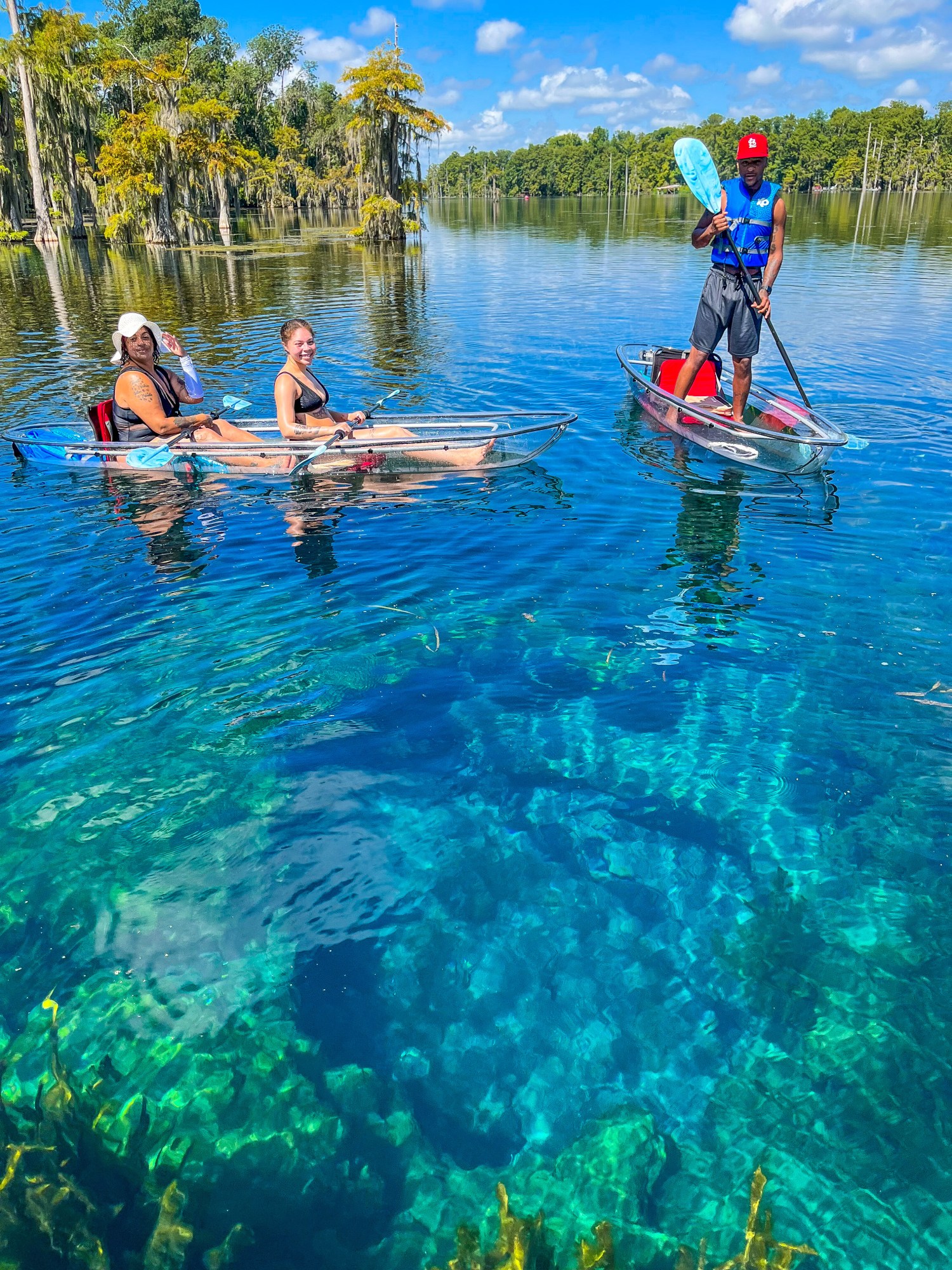People in clear kayaks paddling on a vibrant blue lake under a sunny sky.