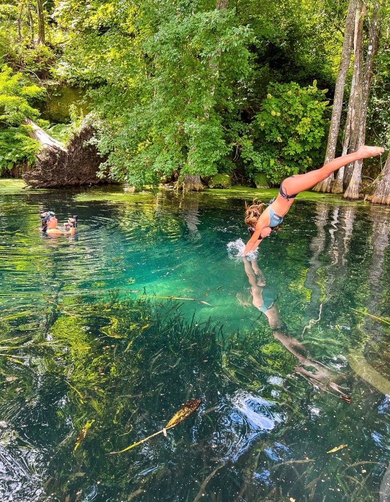 Person diving into a clear, forested river while another swimmer takes a picture.