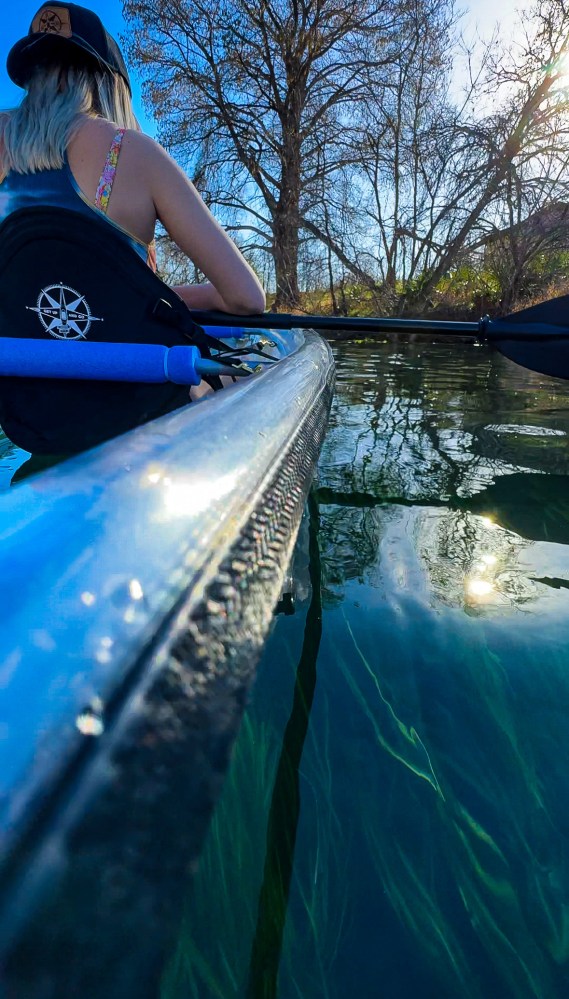 Person kayaking on clear water with visible aquatic plants and leafless trees in the background.