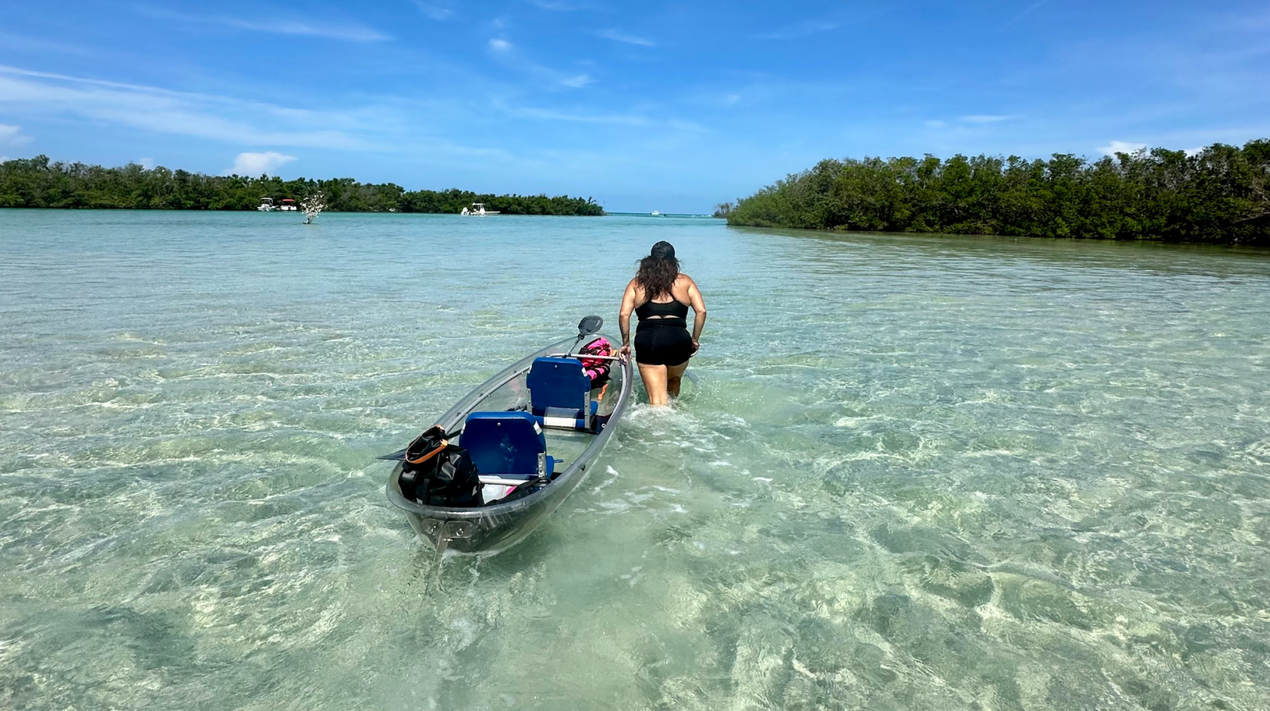 Woman in swimsuit pulls transparent kayak through shallow clear water near mangroves.