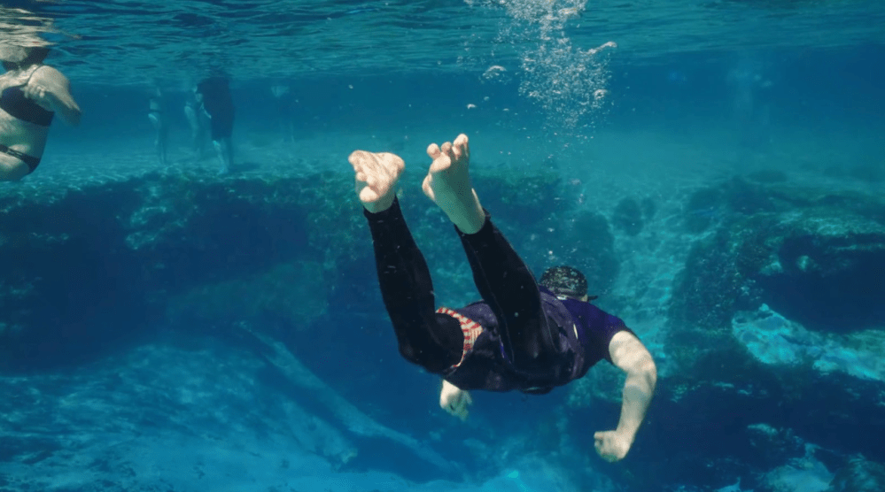 Person snorkeling underwater in clear blue sea near rocks.