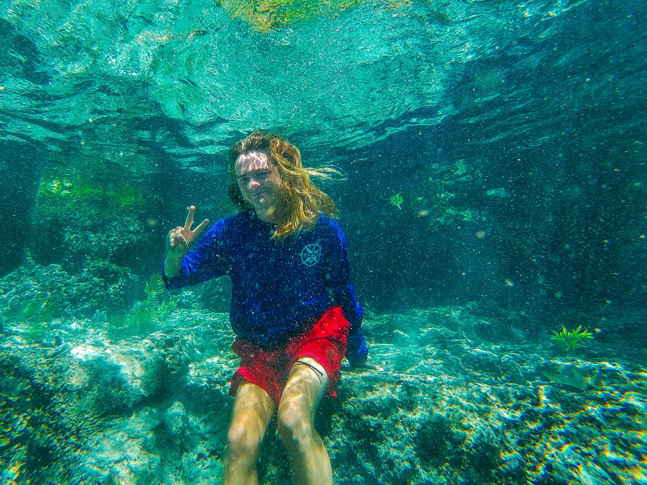 Person underwater wearing blue shirt and red shorts, making a peace sign with one hand.