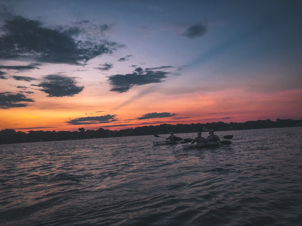 Three kayakers on a lake during a colorful sunset with clouds.