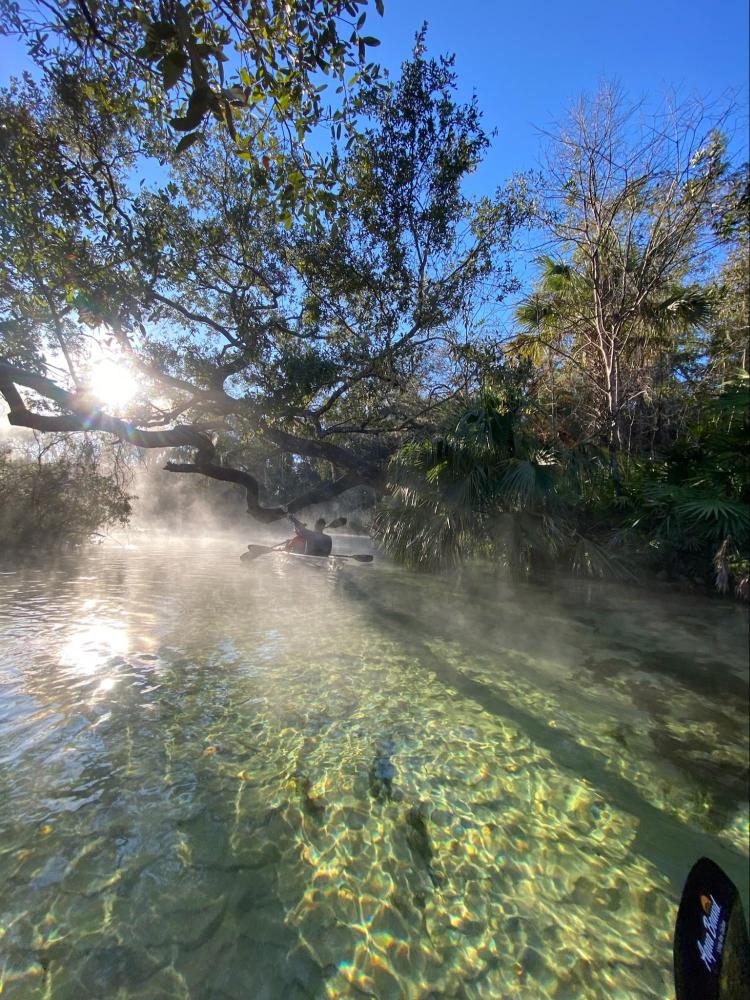 Kayaker on clear, sunlit river with trees and mist in the background.