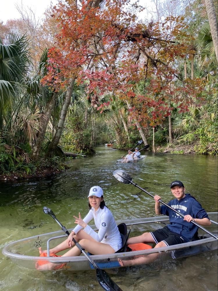 Two people kayaking on a clear river with autumn trees and greenery around.