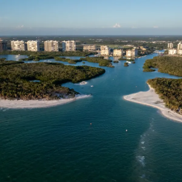 Aerial view of a coastal inlet with sandy beaches and distant buildings under a clear sky.