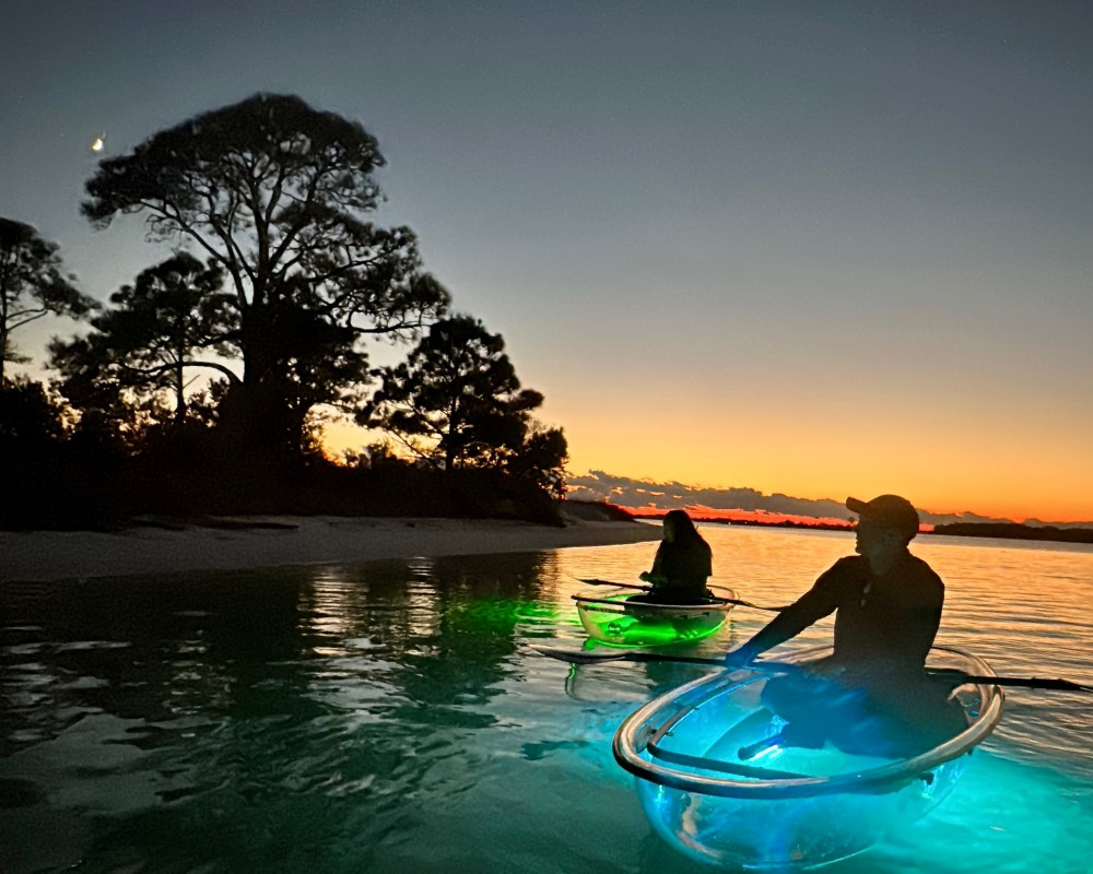 Two people kayaking in glowing clear kayaks at sunset near trees.