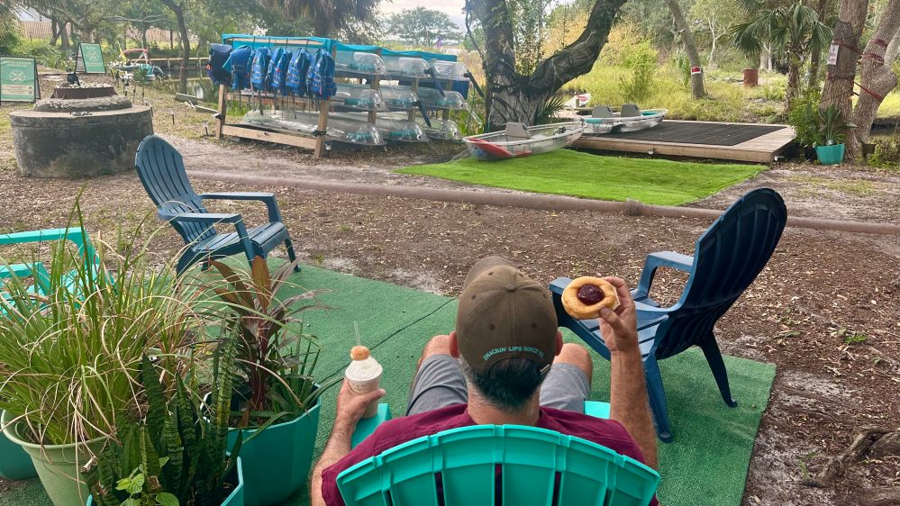 Man in chair holds donut and drink, facing boats and life jackets in outdoor setting.