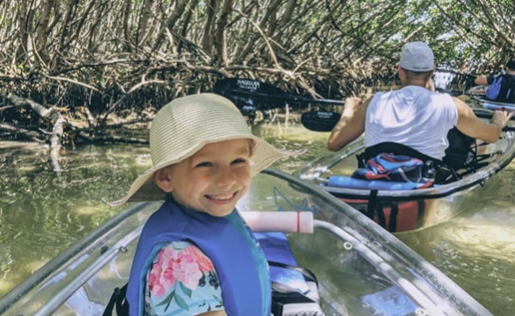 Smiling child in a hat kayaking through mangroves with an adult in another kayak nearby.
