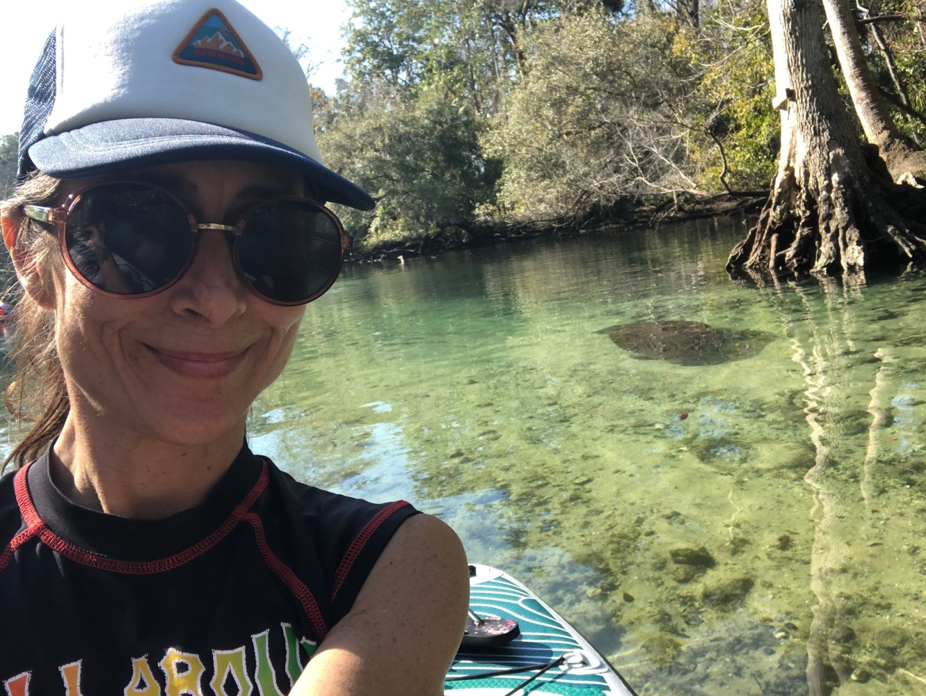 Woman in hat and sunglasses smiles on a paddleboard in a clear, tree-lined river.