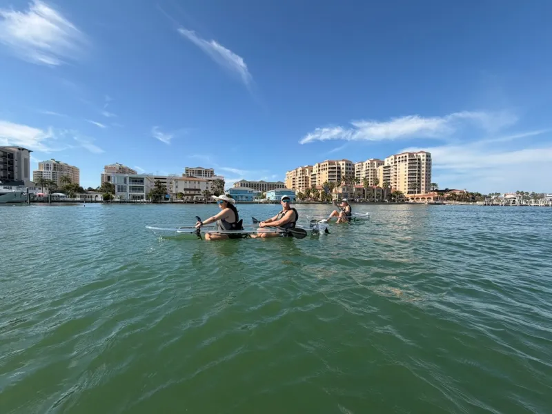 Three people kayaking on calm water with buildings in the background under a clear blue sky.