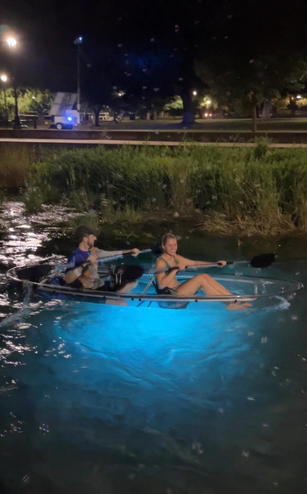 Two people in a clear kayak with blue light paddling on a river at night.