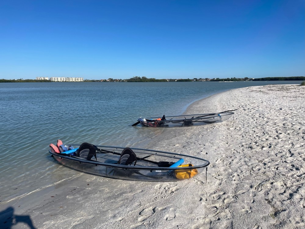 Two clear kayaks on a sandy beach by a calm blue sea under a clear sky.