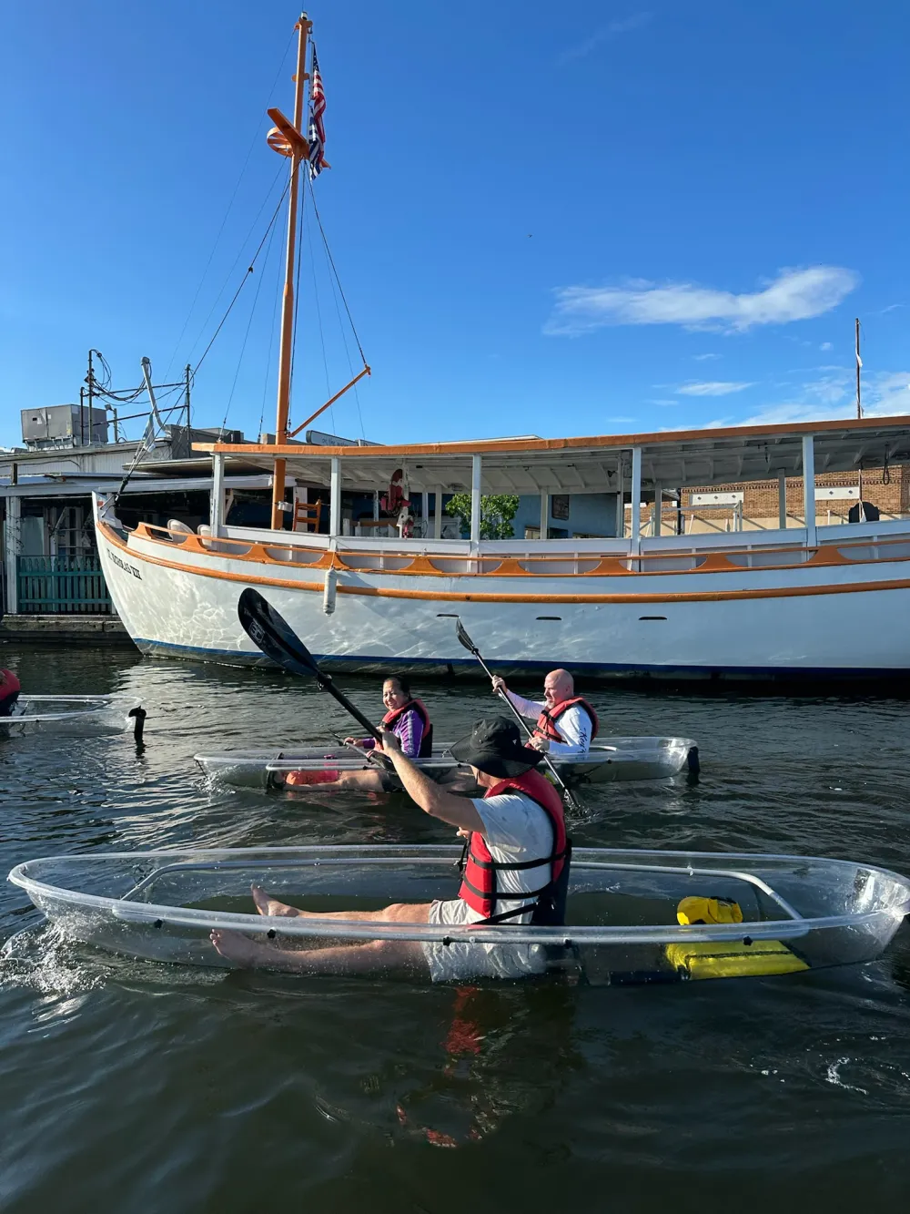 Three people in clear kayaks paddling near a white boat under a bright blue sky.