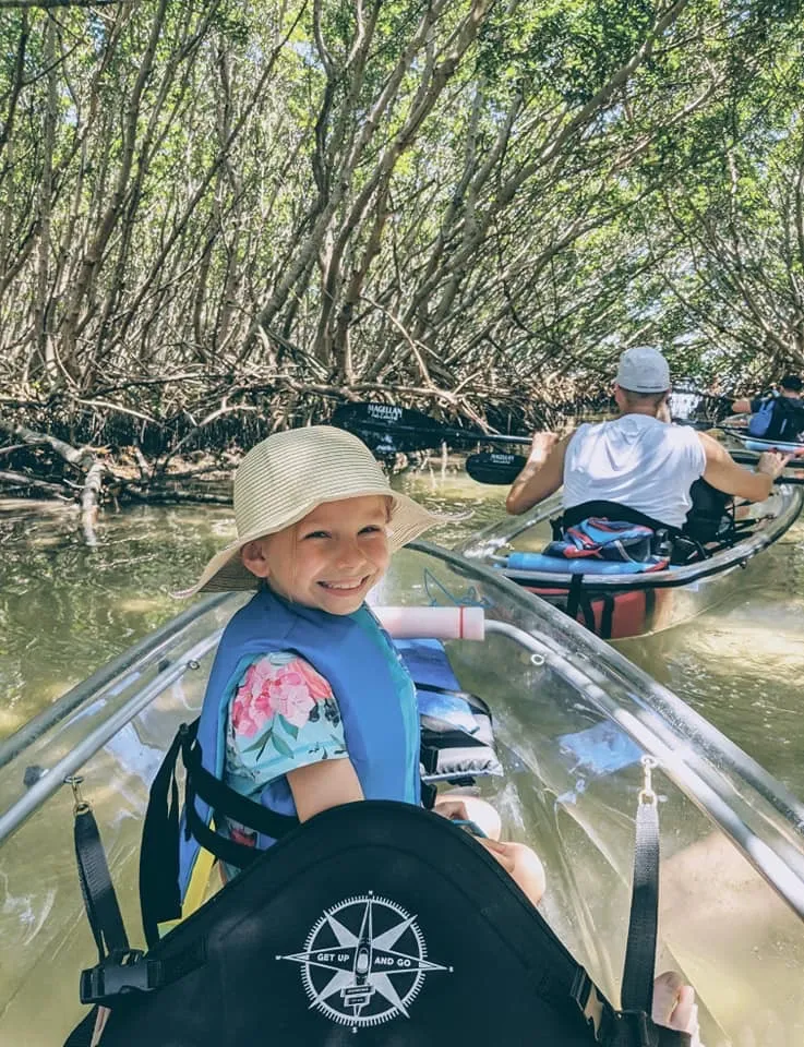 Child in a hat smiles while kayaking through a mangrove tunnel.