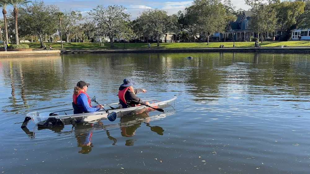 Two people in a transparent kayak paddling on a calm lake near a park with trees and houses.