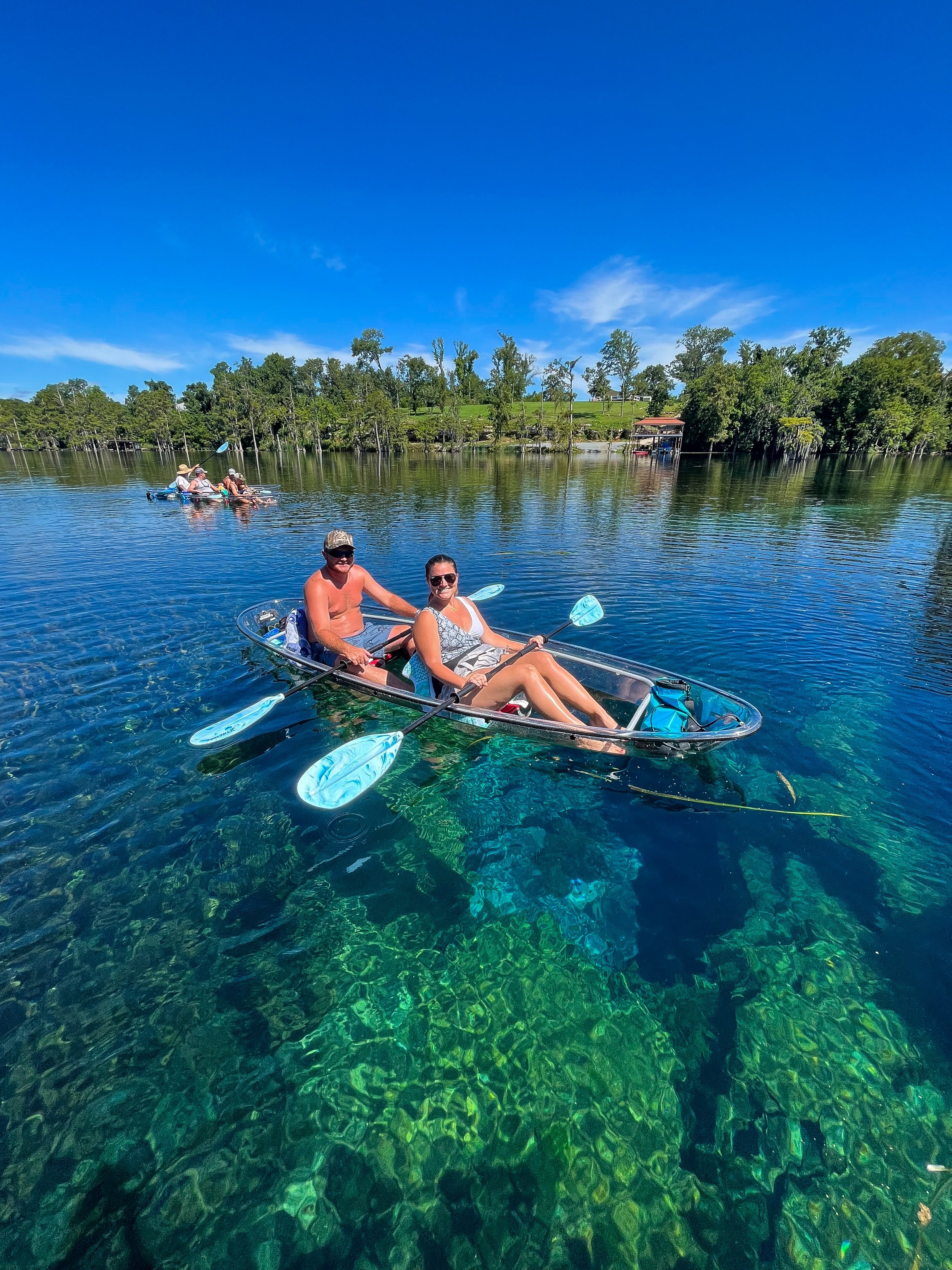Two people in a clear kayak on a blue lake with trees and a clear sky.