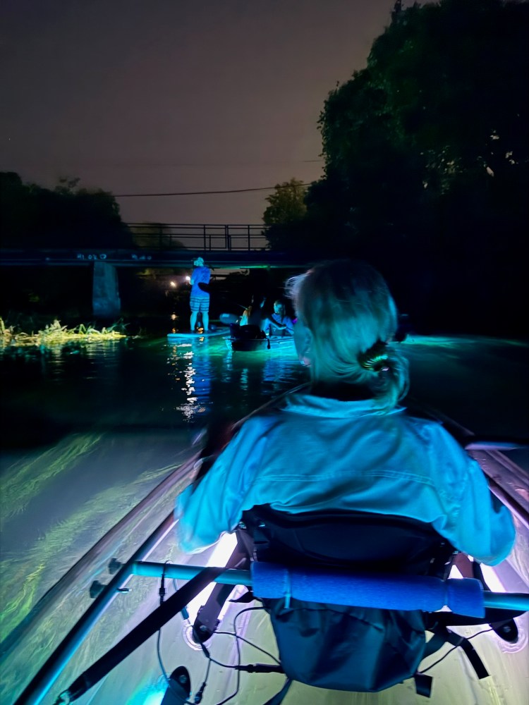 Two people in a clear kayak with blue light underneath, paddling on dark water.