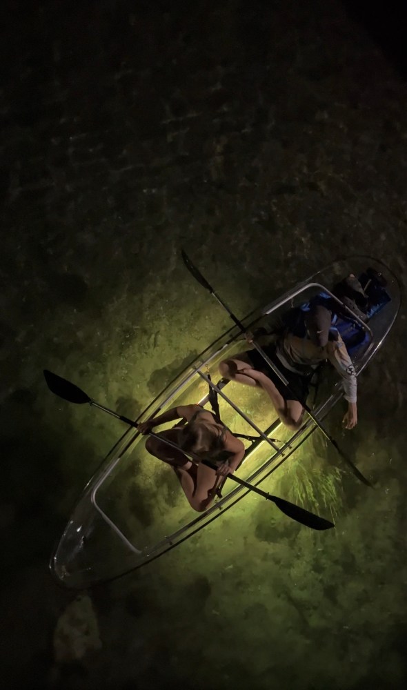 Two people kayaking in a clear kayak over illuminated water at night.