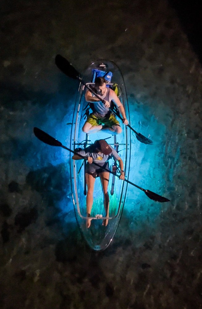 Aerial view of two people kayaking in a transparent canoe with blue light.