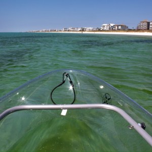 View from transparent kayak on green water with a beach and buildings in the distance.