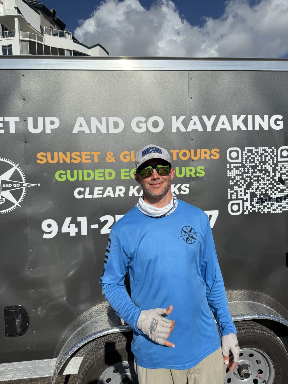 Person in blue shirt and cap stands in front of a kayaking tour trailer smiling.