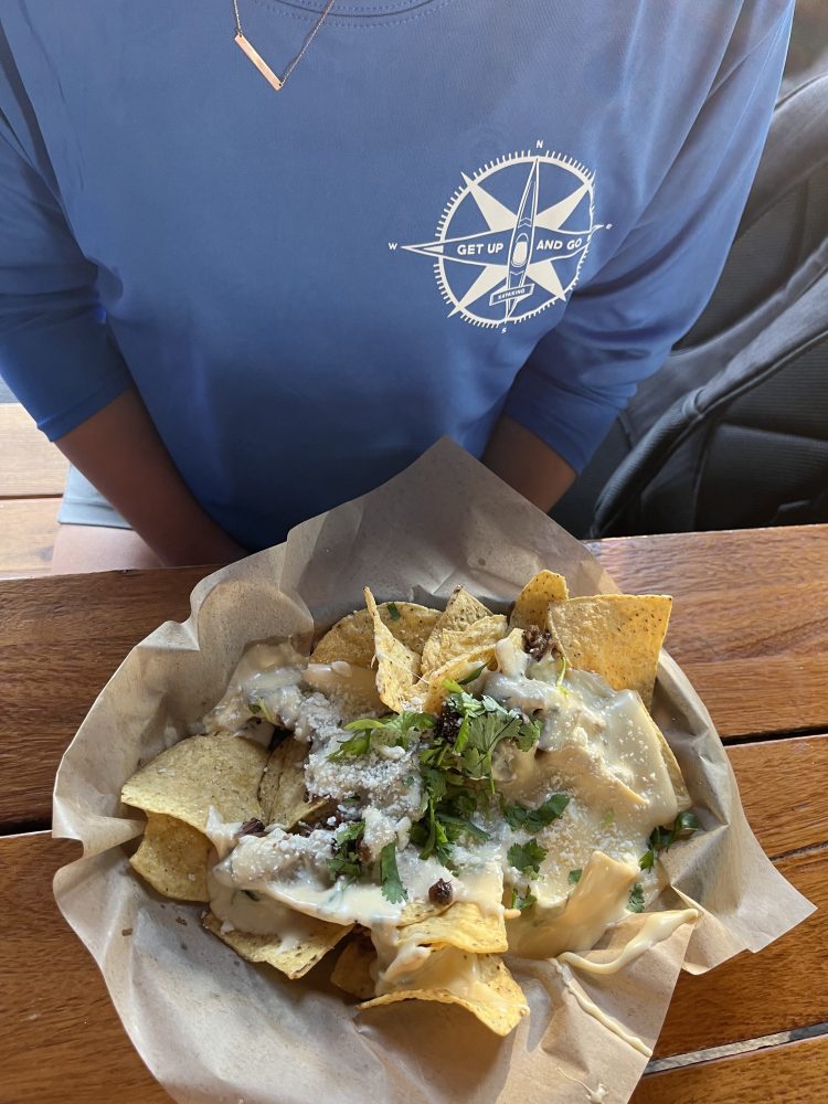 Person in blue shirt sitting at table with a bowl of cheesy nachos.