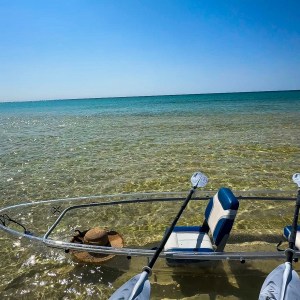 Clear kayak with paddles on shallow, sandy beach under a blue sky.
