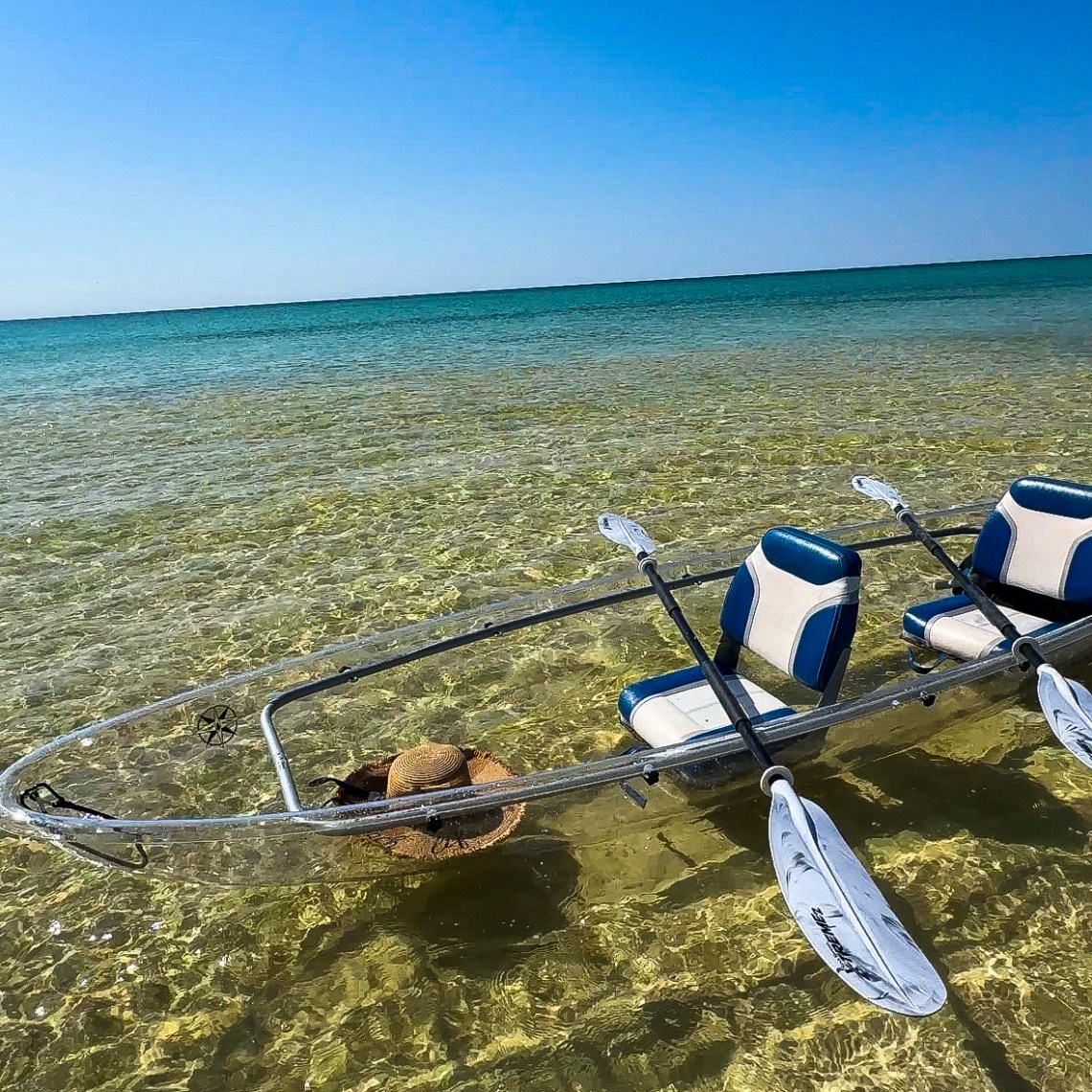 Clear kayak with two blue seats floats on shallow, clear water under a blue sky.