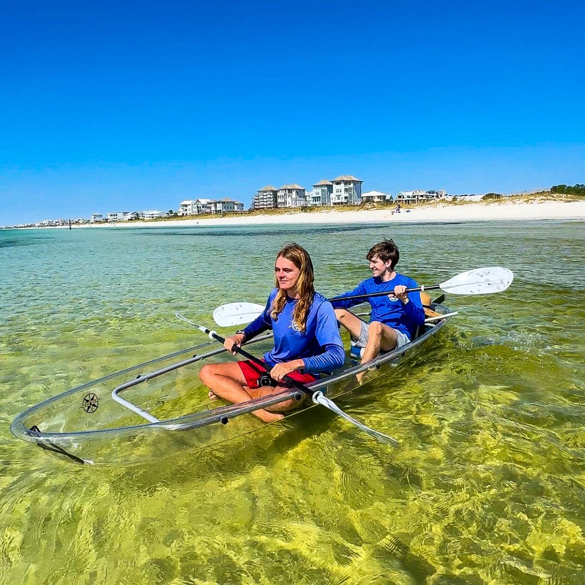 Two people kayaking in clear water near a sandy beach with buildings in the background.