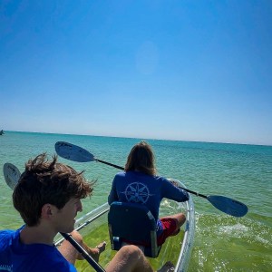 Two people in a clear kayak paddling on a sunny day with a bright blue sky and calm water.