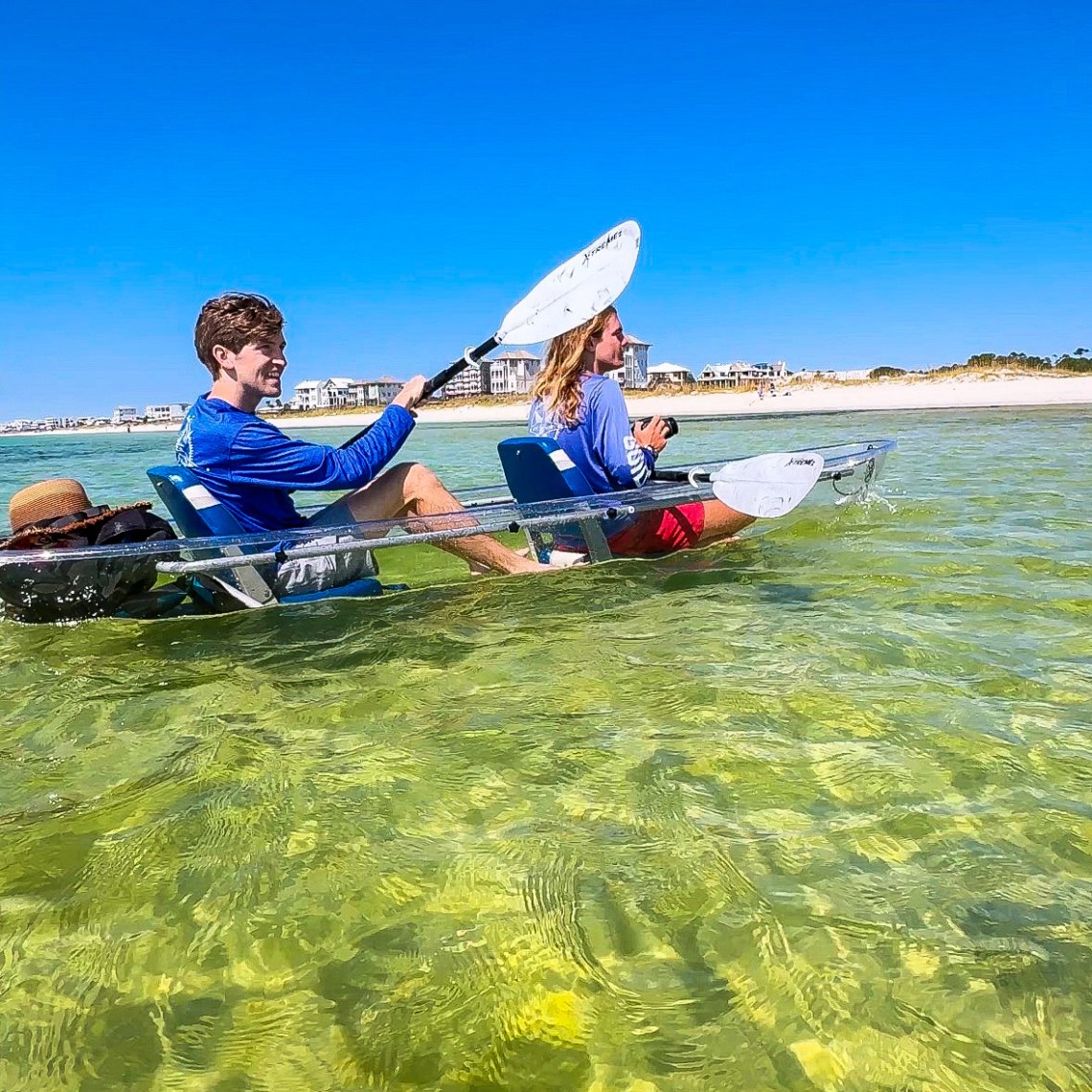 Two people kayaking in a transparent kayak on clear, shallow green water near a sandy shore.