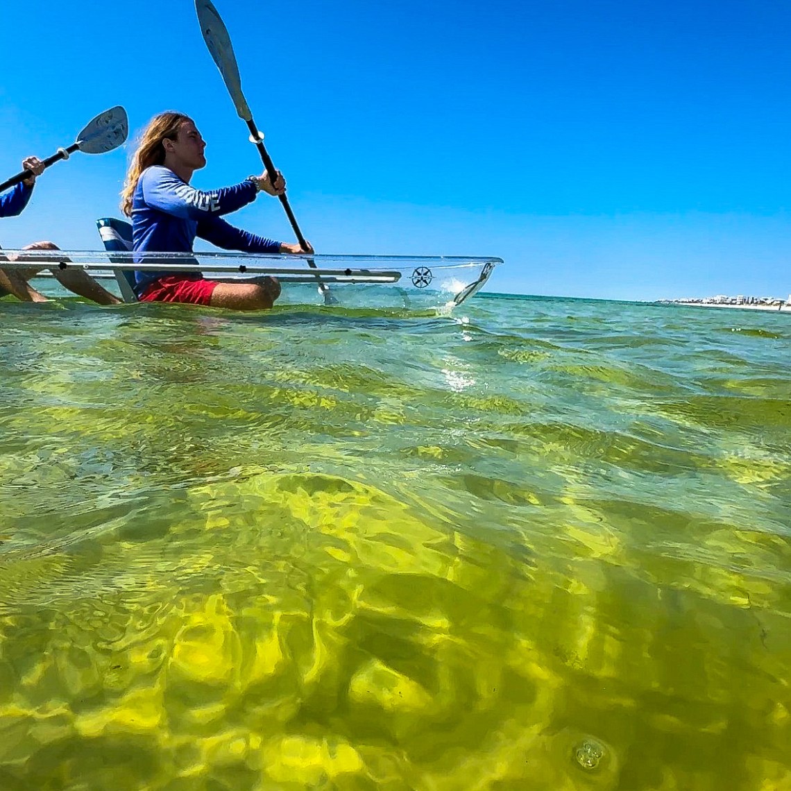 Two people in a clear kayak paddling on greenish water under a blue sky.