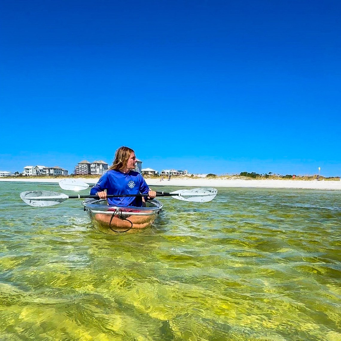 Person kayaking in clear water with buildings and sandy beach in background.