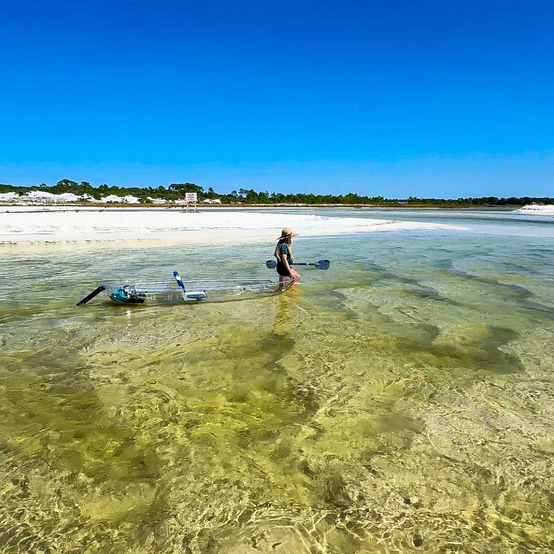 Person with a hat pulling a transparent kayak through shallow, clear water on a sunny day.