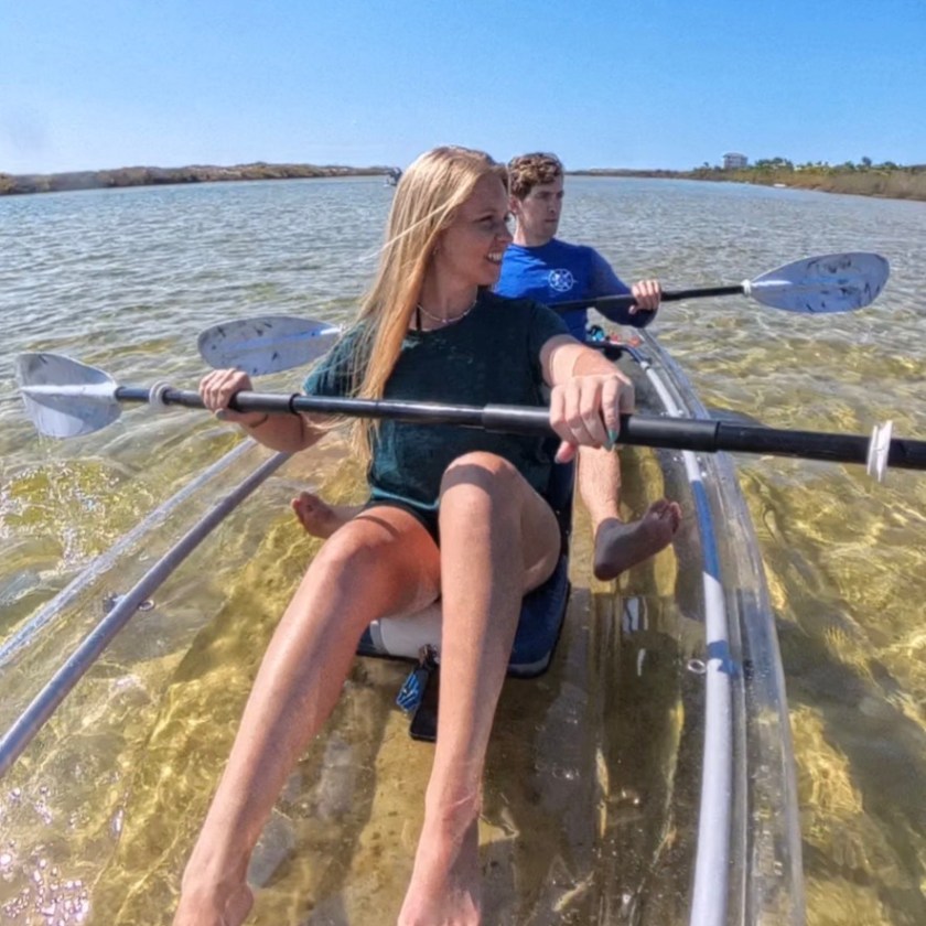 Two people kayaking on a clear kayak in shallow water under a blue sky.