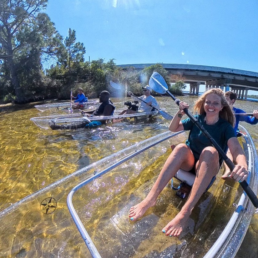 People kayaking in transparent canoes on a sunny day, with a bridge and trees in the background.