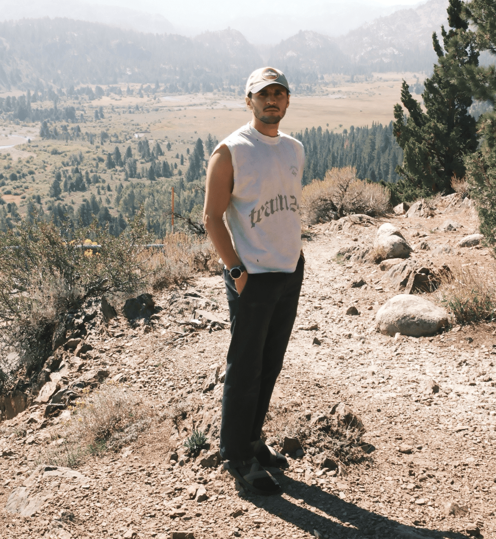 Person in sleeveless shirt and cap standing on rocky trail with mountains in background.