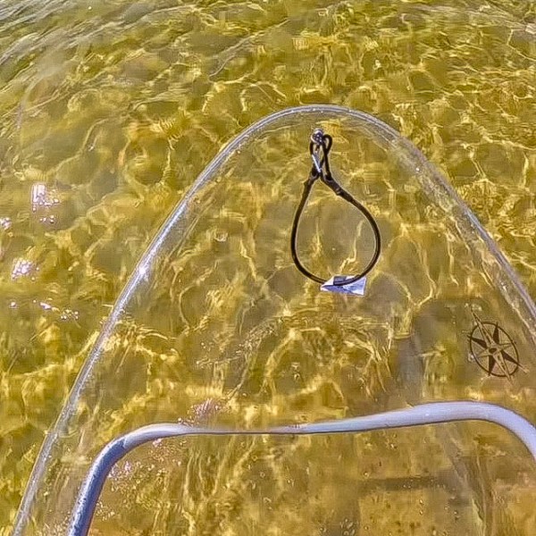 Clear kayak with feet visible over shallow water near a sandy shoreline.