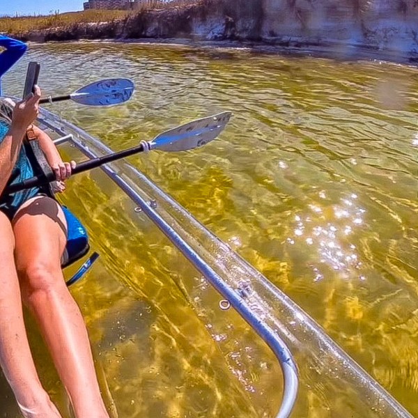 Person kayaking on clear water with legs visible, and grass on a sandy bank under a blue sky.